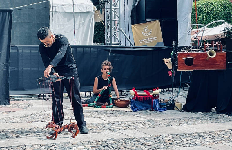 Person handling dog puppet while another plays a snake-shaped flute