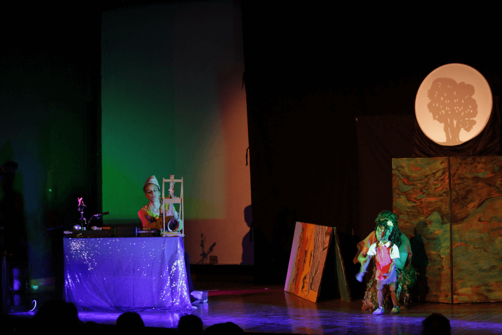 Zuuk plays the self-built ngoni harp while Iracema manipulates the More puppet. In the background a shadowy tree is projected.